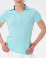 Photo d'un polo de golf bleu ciel à col baseball et boutonné porté par une jeune femme les mains sur les hanches avec un bas blanc et le tout sur fond gris très clair