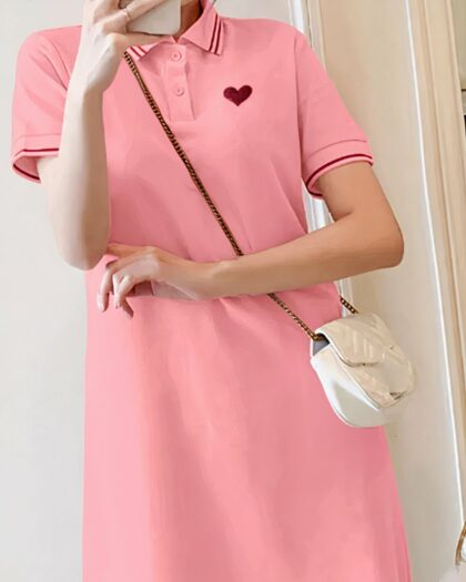 Photo d'une jeune femme debout devant un mur blanc portant une robe polo rose boutonnée avec un coeur brodé rouge sur le côté gauche