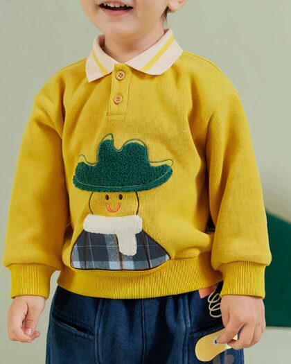 On voit un petit garçon habillé d'un polo manches longues jaune avec un personnage.