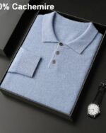 Photo d'un pull polo bleu plié dans une boîte noire ouverte posée sur un socle gris foncé une montre d'homme posée à côté et l'inscription 100% cachemire en haut à gauche