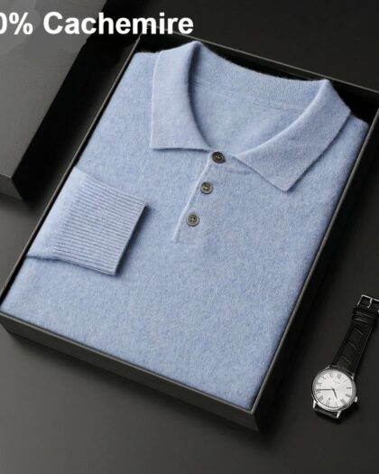 Photo d'un pull polo bleu plié dans une boîte noire ouverte posée sur un socle gris foncé une montre d'homme posée à côté et l'inscription 100% cachemire en haut à gauche