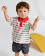 On voit un petit garçon brun qui porte un polo blanc et rouge avec un short cargo gris foncé