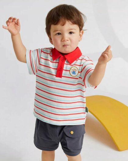 On voit un petit garçon brun qui porte un polo blanc et rouge avec un short cargo gris foncé
