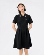 Photo d'une jeune femme debout les mains dans le dos portant une robe polo noire à col rayé noir et blanc le tout sur un fond bleu pâle