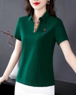 Photo d'une femme portant un polo de golf vert foncé avec petit décolleté en V en tissu Burberry avec un petit écusson un homme tapant une balle au golf : elle est appuyée avec les bras tendus sur un meuble et sur un fond foncés