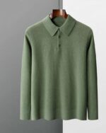 Photo d'un pull polo chandail vert sur un cintre en bois marron foncé manches à la même hauteur que le bas du pull le tout sur un fond gris foncé à gauche et bleu pâle à droite