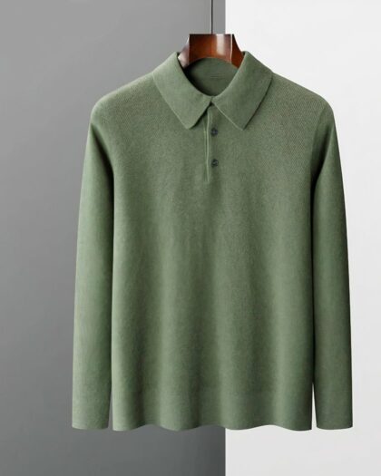 Photo d'un pull polo chandail vert sur un cintre en bois marron foncé manches à la même hauteur que le bas du pull le tout sur un fond gris foncé à gauche et bleu pâle à droite