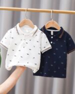 Sur une tringle, on voit deux polos pour bébé garçon, un blanc et un bleu marine. Les deux sont imprimés de dinosaures.