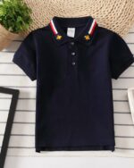 On voit un polo bleu marine pour bébé garçon sur un fond en lambris blanc, près d'une plante verte