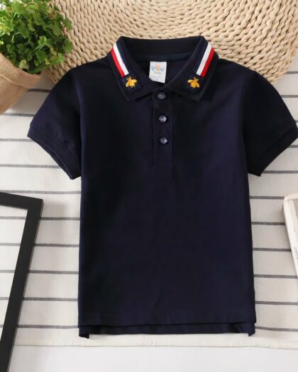 On voit un polo bleu marine pour bébé garçon sur un fond en lambris blanc, près d'une plante verte