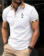 Photo d'un homme adossé à un mur de verre noir les mains dans les poches d'un pantalon beige et portant un polo de golf blanc avec une lettre noire sur la poitrine et des ornements à carreaux autour des manches courtes, au col et à la patte de boutonnage