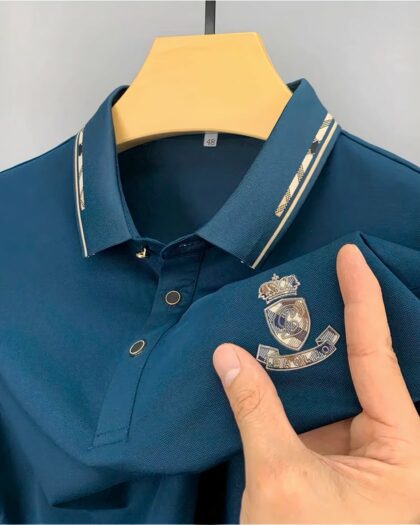 Photo représentant un polo bleu à écusson tenu par une main masculine et pendu sur un cintre an bois sur un fond gris