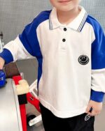 On voit un enfant qui porte un polo manches longues blanc et bleu avec un écusson smiley.