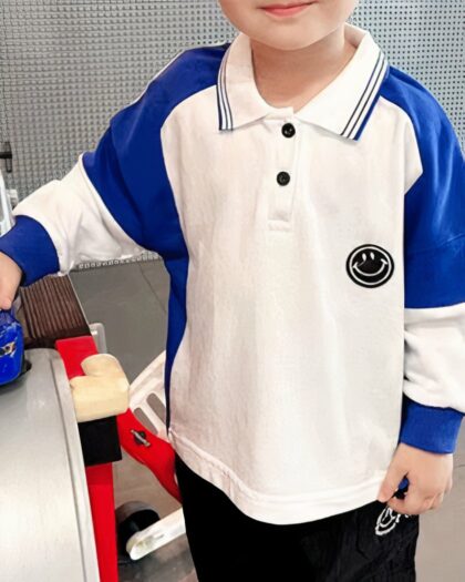 On voit un enfant qui porte un polo manches longues blanc et bleu avec un écusson smiley.