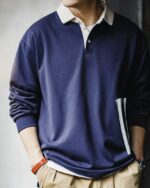 Photo d'un homme portant un polo vintage à manches longues bleu marine avec un livre sous le bras et les mains dans les poches de son pantalon beige.