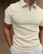Polo zippé uni beige porté par un homme avec un pantalon blanc, les mains dans les poches.
