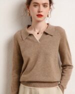 Photo d'une femme adossée à un mur beige portant un pull polo marron clair en cachemire sans bouton et la main gauche dans la poche d'un pantalon beige