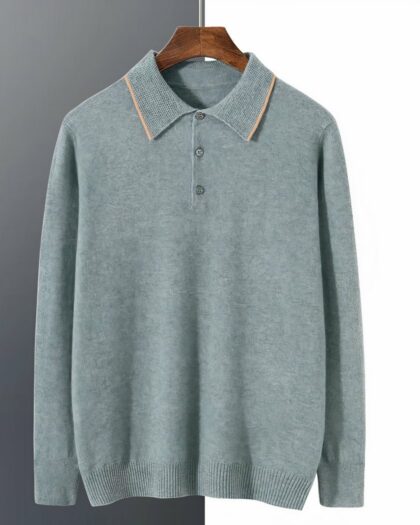 Photo représentant un pull polo bleu en cachemire à col bicolore et petits boutons sur un cintre en bois marron le tout devant un mur blanc à droite et gris foncé à gauche