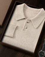 Photo d'un pull polo beige en cachemire plié dans une boîte ouverte avec son col polo et patte de boutonnage et deux boutons une montre est posée à sa droite