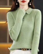 Photo d'une jeune femme brune debout devant une étagère jaune et métal d'un côté et un fond noir de l'autre une main sur son cou et portant un pull polo vert en laine mélangée et un bas blanc avec une ceinture foncée à boucle dorée