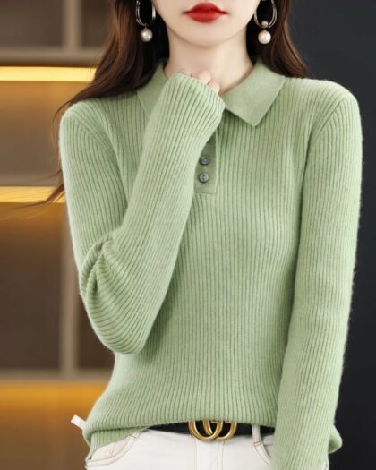 Photo d'une jeune femme brune debout devant une étagère jaune et métal d'un côté et un fond noir de l'autre une main sur son cou et portant un pull polo vert en laine mélangée et un bas blanc avec une ceinture foncée à boucle dorée