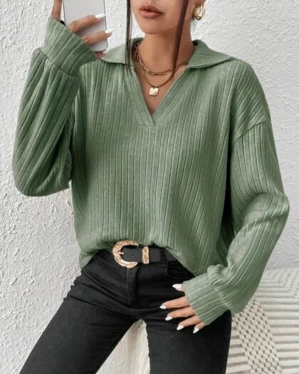 Photo d'une femme debout téléphone en main droite et main gauche sur poche du jean noir à ceinture à boucle, portant un pull polo vert à manches longues col large et encolure en V le tout devant un mur blanc