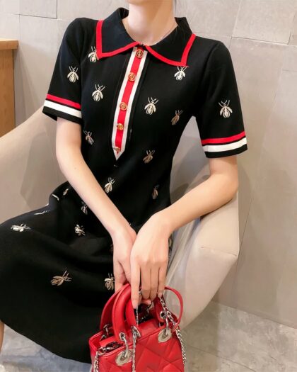 Photo d'une jeune femme assise dans un fauteuil blanc et portant une robe polo noire à manches courtes imprimé insectes ailés blancs bord de col rouge, bords de manches rouge et blanc et patte de boutonnage rouge et blanche à boutons dorés avec un sac rouge dans les mains