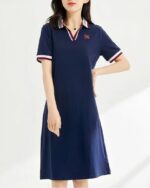 Photo d'une jeune femme debout une main à la taille portant une robe polo bleu marine avec col, décolleté et bord des manches tricolores le tout devant un mur bleu pâle à rays de lumière blanche