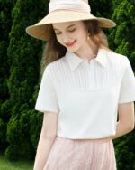Photo représentant une jeune femme aux cheveux longs debout bras gauche plié dans le dos et bras droit le long du corps portant une capeline en paille, un polo blanc en coton à empiècement arrondi devant et une jupe rose devant un massif d'arbres vert foncé