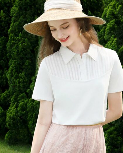 Photo représentant une jeune femme aux cheveux longs debout bras gauche plié dans le dos et bras droit le long du corps portant une capeline en paille, un polo blanc en coton à empiècement arrondi devant et une jupe rose devant un massif d'arbres vert foncé