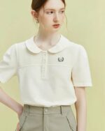 Photo d'une jeune femme main droite sur la hanche portant un polo chic blanc à col claudine et petite broderie, manches légèrement bouffantes rentré dans un bas kaki le tout sur un fond vert clair