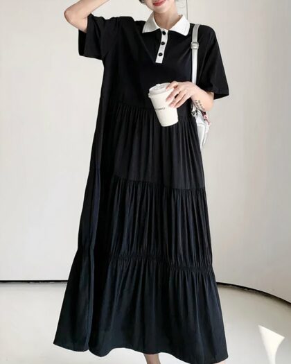 Photo d'une robe polo noire au col et patte de boutonnage blanche longue et ample portée par une jeune femme tenant un gobelet dans la main gauche un sac en bandoulière blanc sur l'épaule gauche et la main droite dans les cheveux, le tout sur un fond gris clair