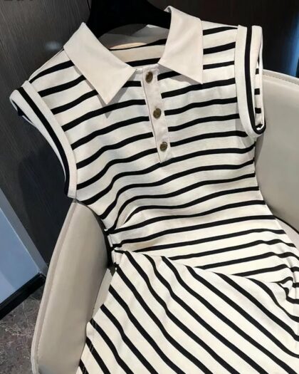 Photo d'une robe polo rayée noire et blanche pour femme sans manche avec un col blanc et des boutons noirs sur patte de boutonnage blanche, la robe sur cintre noir est posée sur un fauteuil gris clair devant une porte en bois gris et un mur bleu