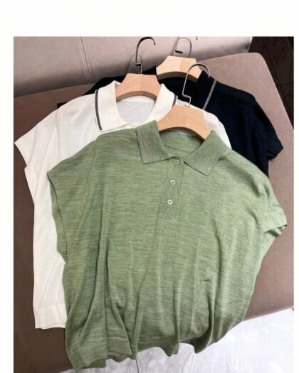 Photo de trois polos à col ornés de petites perles sur cintre posés les uns sur les autres en léger décalé, un gris foncé en dessous, un blanc au milieu et un vert dessus polos à emmanchures larges et plates formant des manches tombantes en tissu souple le tout sur un fond dégradé de gris