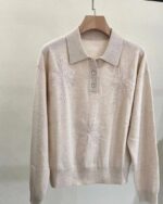 Photo d'un pull polo rose pâle côtes bas et bas des manches, broderies avec strass sur un cintre en bois le tout sur fond gris