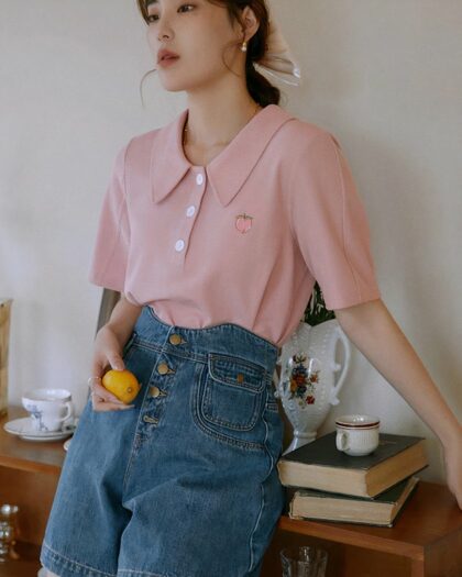 Photo d'une femme portant un polo vintage ample rose, brodé à large col avec un jean taille haute, appuyé sur un meuble en bois avec une orange dans la main.