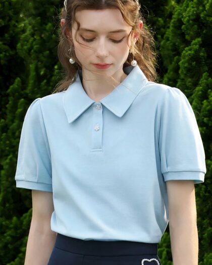 Photo d'une jeune femme debout portant un polo chic bleu à manches courtes ballon, col polo et deux boutons blancs sur un bas bleu marine avec un ornement coeur argenté sur le c$^té le tout devant un massif d'arbres vert foncé