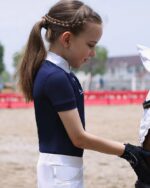 Une petite fille habillée aevc un polo blei et un pantalon blanc qui caresse la tête d'un cheval