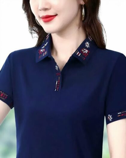 photo du buste d'une femme portant un polo bleu marine orné de broderies de couleur rouge, blanche et marron sur le tour du bas des manches, le col et la patte de boutonnage, le tout sur un fond flou de verdure et de ciel