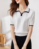 Photo représentant une femme debout tenant son sac à mains devant elle à deux mains et portant un polo chic blanc à liseré noir au bord des manches, tour de col et encolure sur un pantalon noir le tout dans une pièce ensoleillée au mur gris clair et un bout de meuble en bois clair avec un bout de tableau