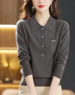 Photo d'une femme debout mains devant portant un pantalon beige et un pull polo gris foncé avec une poche poitrine ornée d'un petit écusson clair le tout sur un fond éclairage doré sur la gaucheet noir à droite