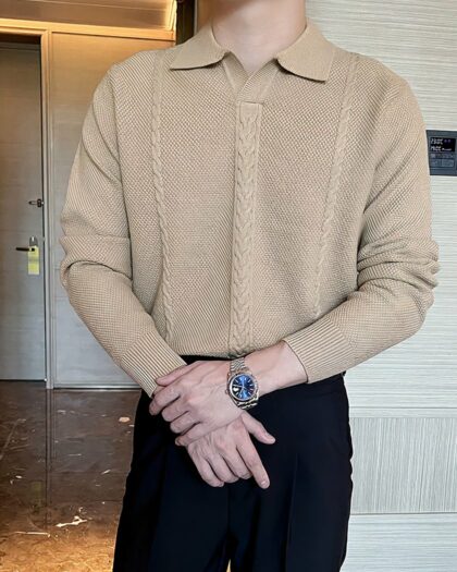 Photo d'un homme debout dans une pièce au mur gris à gauche, au fond une porte grise et un sol marron, les mains croisées devant portant un polo beige sans bouton avec un torsade milieu devant assez grande et une plus petite de chaque côté