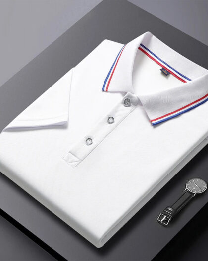 Un polo chic col bleu blanc rouge pour homme blanc plié, avec col et manches bordés de bleu et rouge, posé sur un fond gris avec une montre à côté.